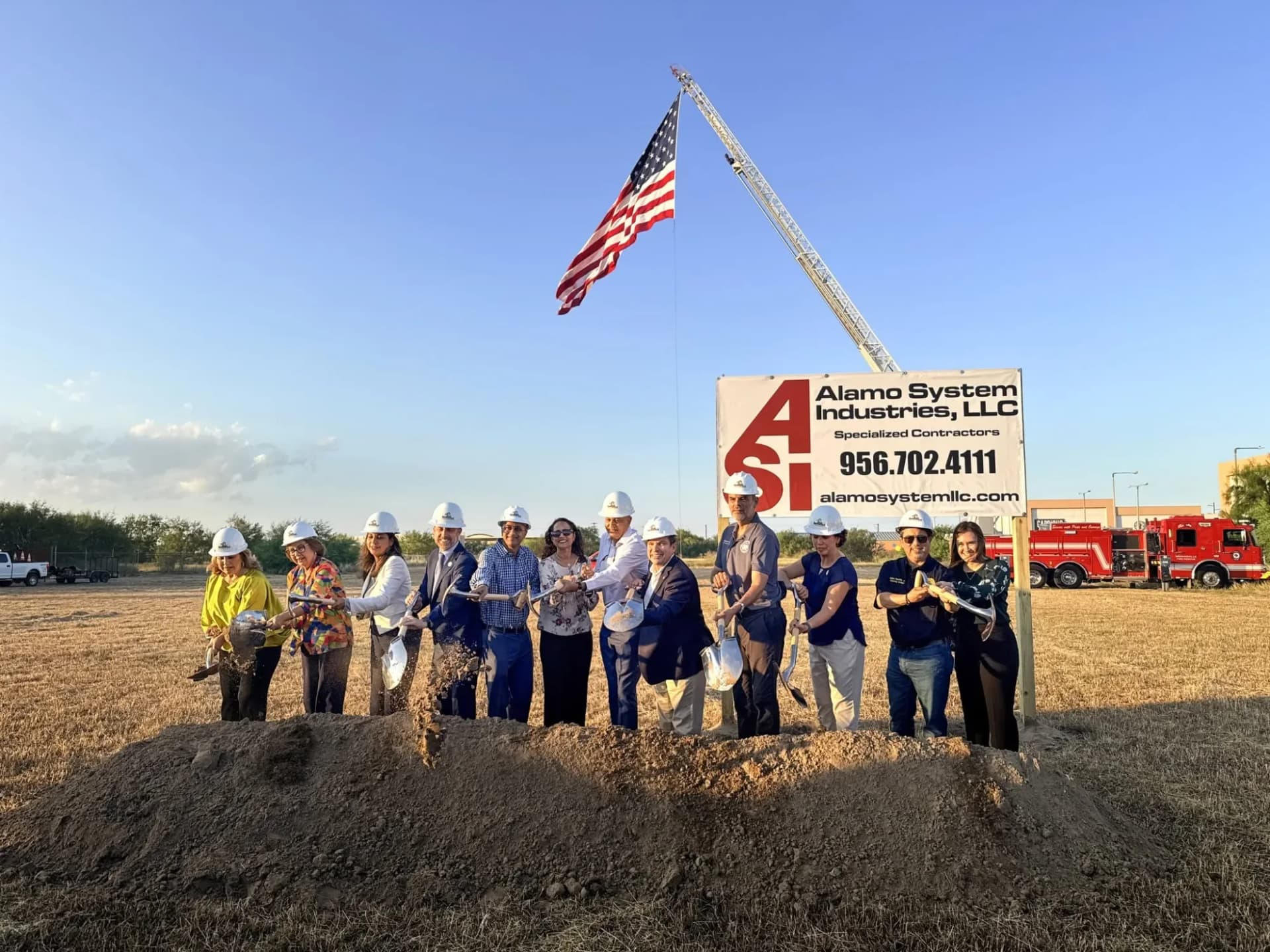 Officials in white hard hats turn dirt with ceremonial shovels at the Embassy Suites by Hilton Brownsville site, under an American flag hung from a fire truck ladder