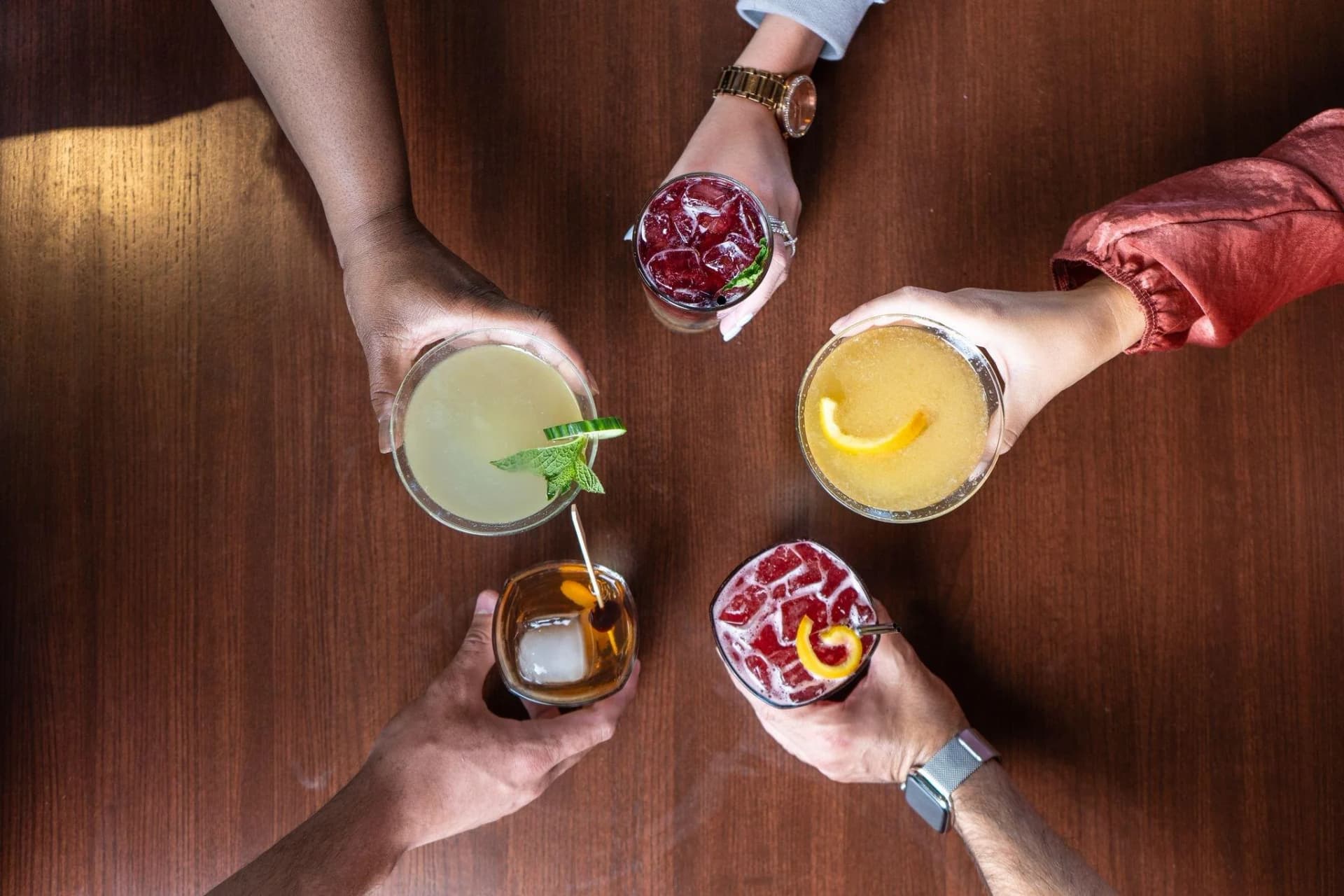 Five cocktails raised in a toast over a wooden table, seen from above