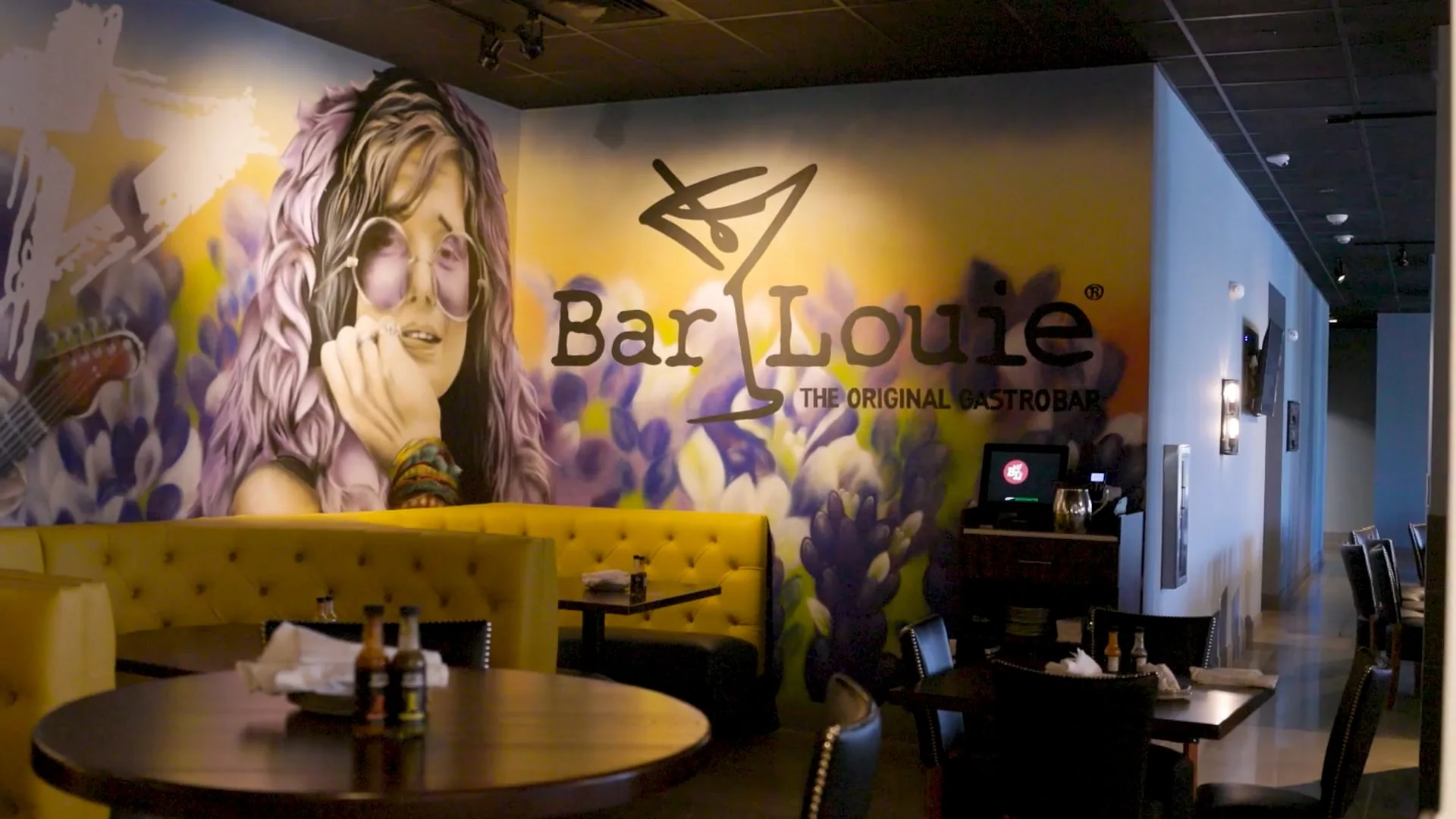 Bar Louie dining room with yellow booths and a mural of a woman in round glasses beside the Bar Louie sign